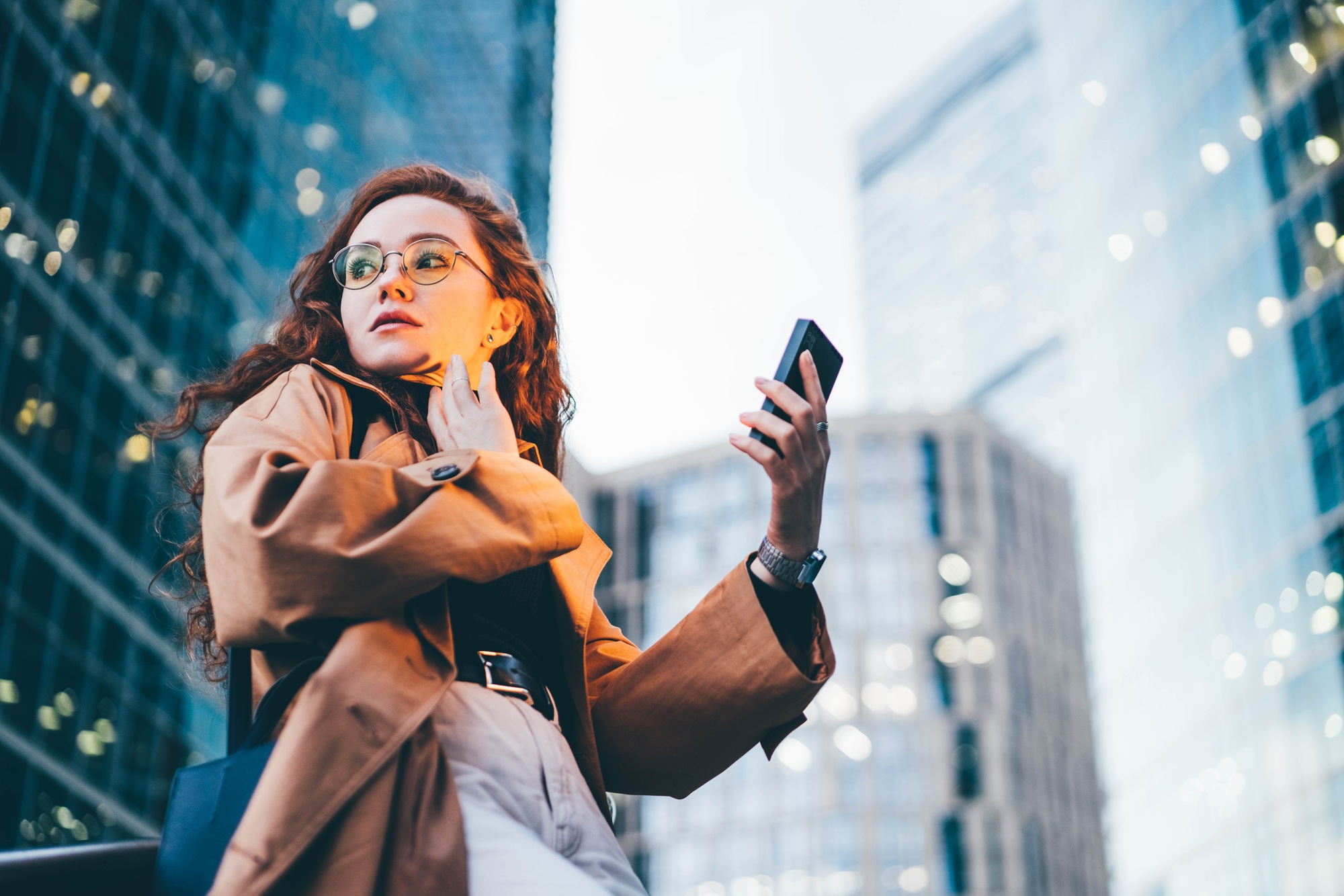 Successful woman using smartphone outdoors while standing near skyscraper at night.